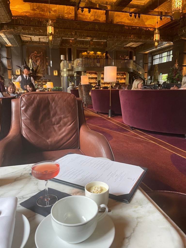 A table set for afternoon tea at ATLAS bar, featuring a pink cocktail, a menu, and a white teacup with a dessert in a small cup, surrounded by an elegant interior decorated with warm colors and plush seating.
