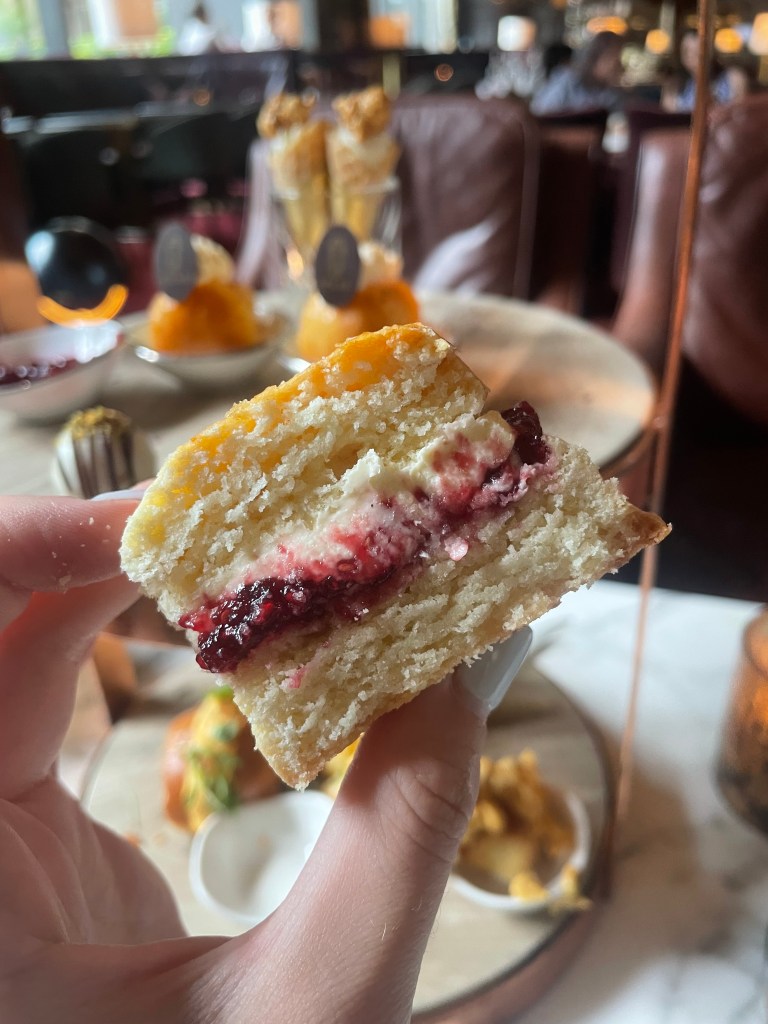 A close-up of a hand holding a half-cut scone filled with cream and jam, with a luxurious café setting in the background.